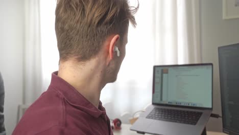 un joven se pone auriculares inalámbricos en los oídos y luego se los quita mientras trabaja en la oficina de su casa