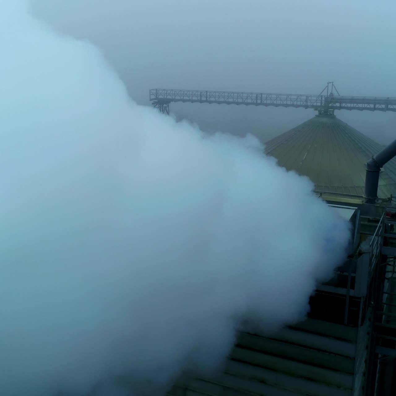 A number of pipes producing thick white smoke at agricultural plant. Rounded roof of silo tank at backdrop. Grey foggy day background