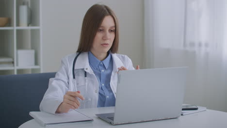 female family doctor is consulting online talking with patient by video chat at laptop working remotely