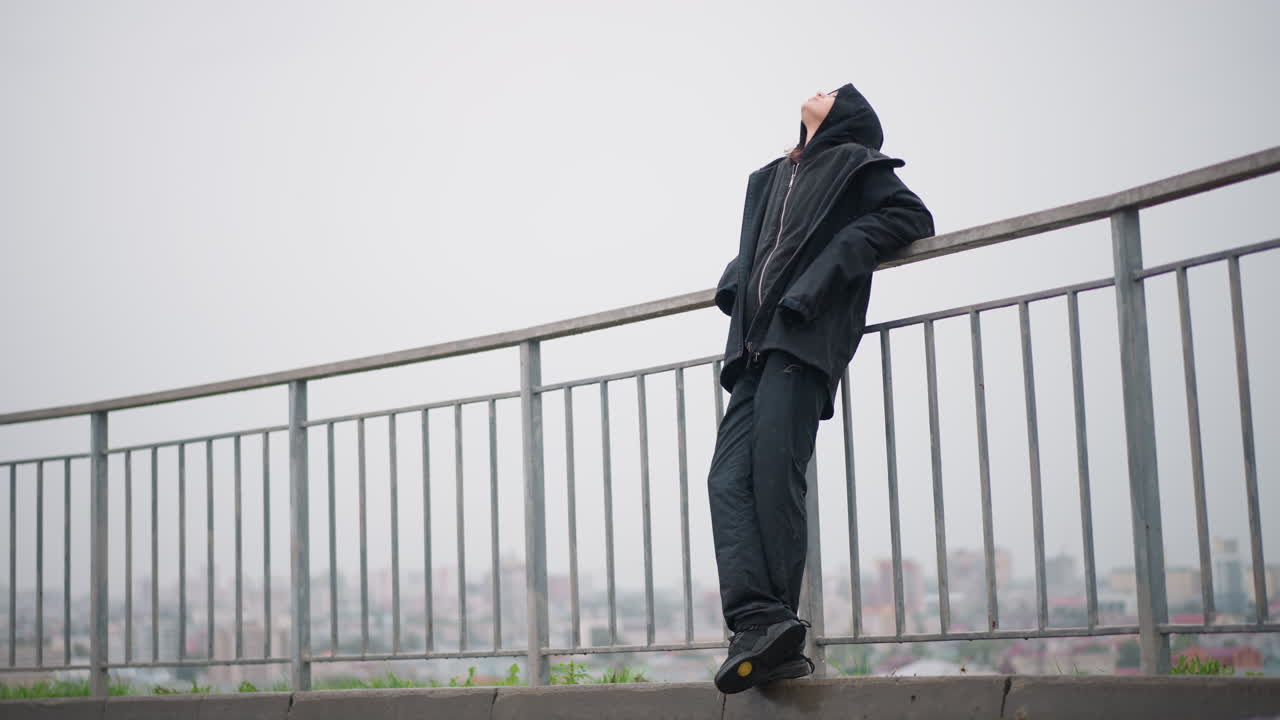 Sollievo nella città piovosa. Una donna fradicia si rilassa appoggiata a una ringhiera all'aperto. Una donna nel paesaggio urbano respira liberamente in una scena urbana sotto la pioggia. Il marciapiede bagnato e lo skyline lontano incorniciano la donna che trova sollievo nella pioggia