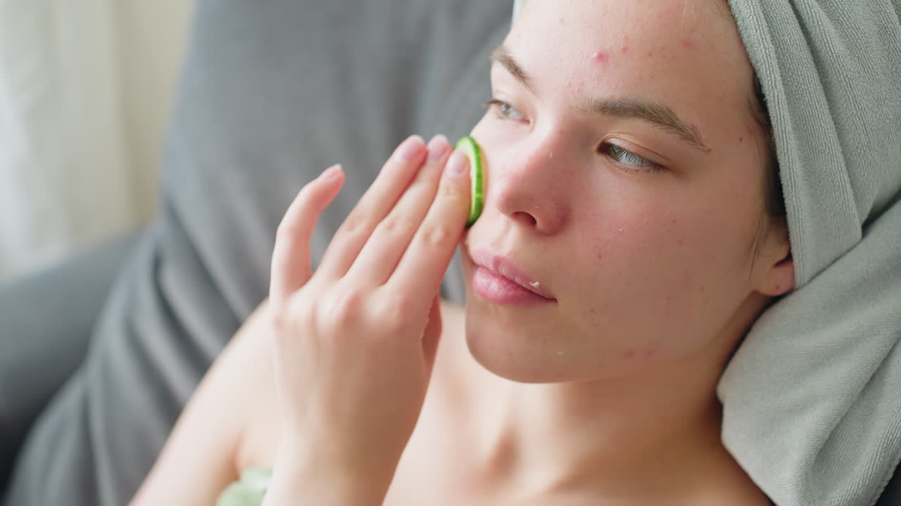 Woman gently rubbing cucumber on face during soothing skincare routine with towel wrapped around hair, sitting in calm indoor space focused on rejuvenation and natural beauty under soft daylight