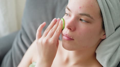 woman gently rubbing cucumber on face during soothing skincare routine with towel wrapped around hair, sitting in calm indoor space focused on rejuvenation and natural beauty under soft daylight