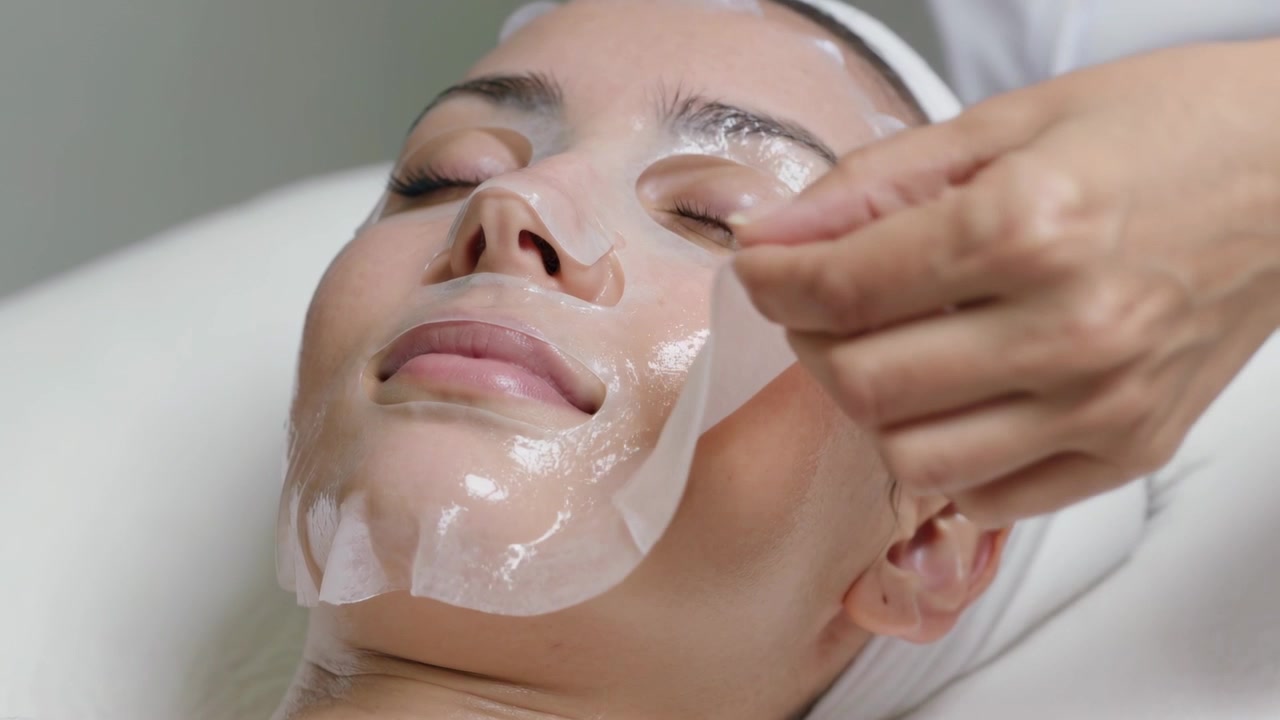 Esteticista de uniforme branco alcançando e levantando máscara de gel da mandíbula na cama, mãos mostrando pele viçosa
