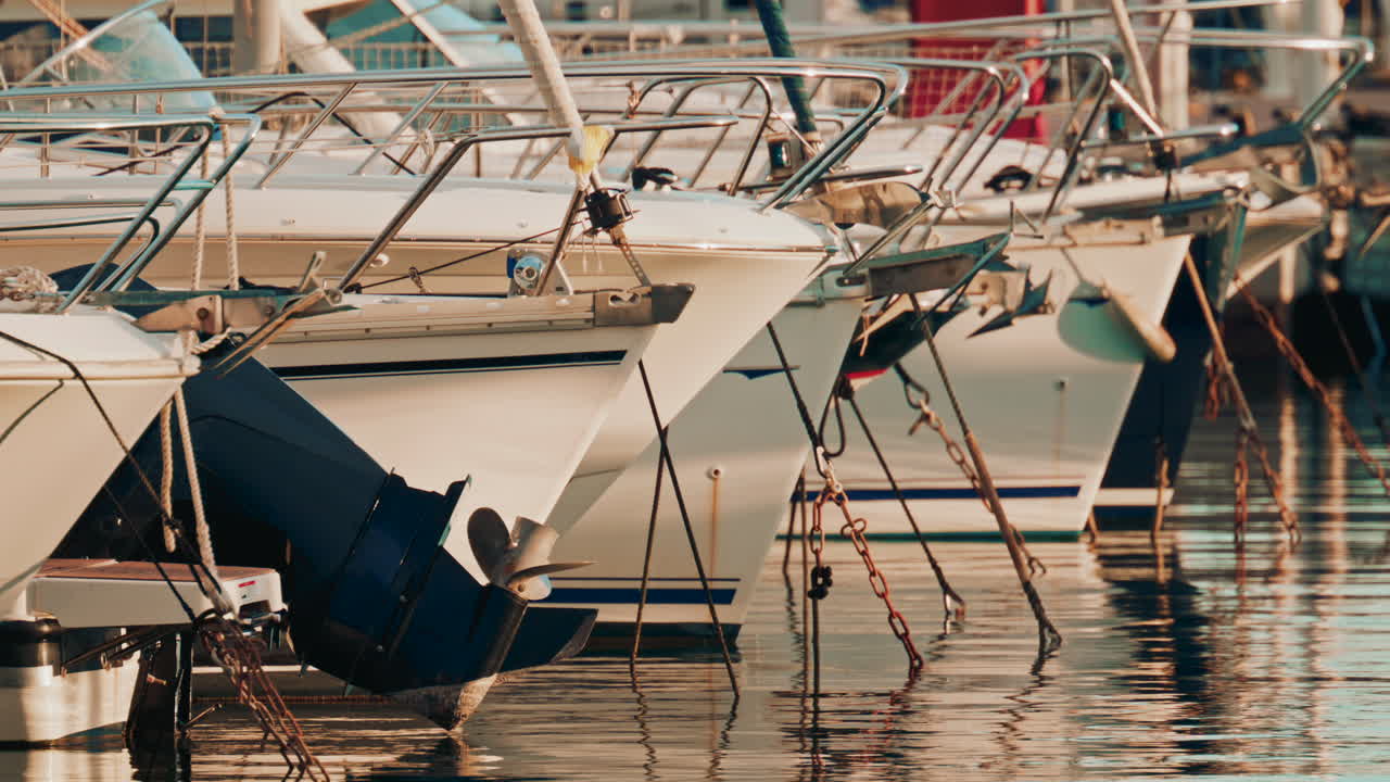 Row of motorboats docked in a marina, showing anchors, railings, and mooring chains in warm sunset light