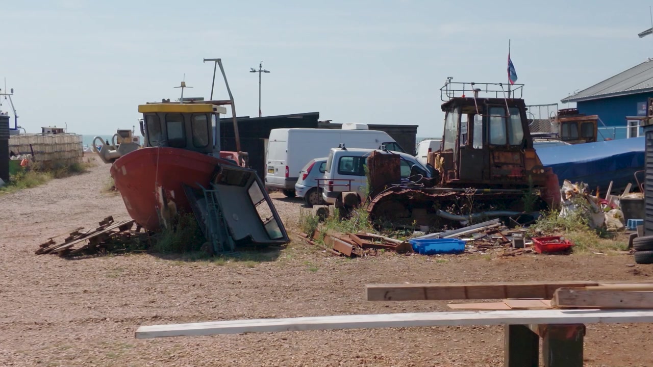 Rød fiskebåd og rusten gravemaskine ligger blandt skrot og maskineri på Hastings strand, England, i et langsomt panorering mod højre.