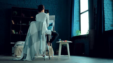 beautiful and young girl paints a picture of oil on canvas in a bright room. easel stands in the studio, paints on the table. circular motion of the camera.