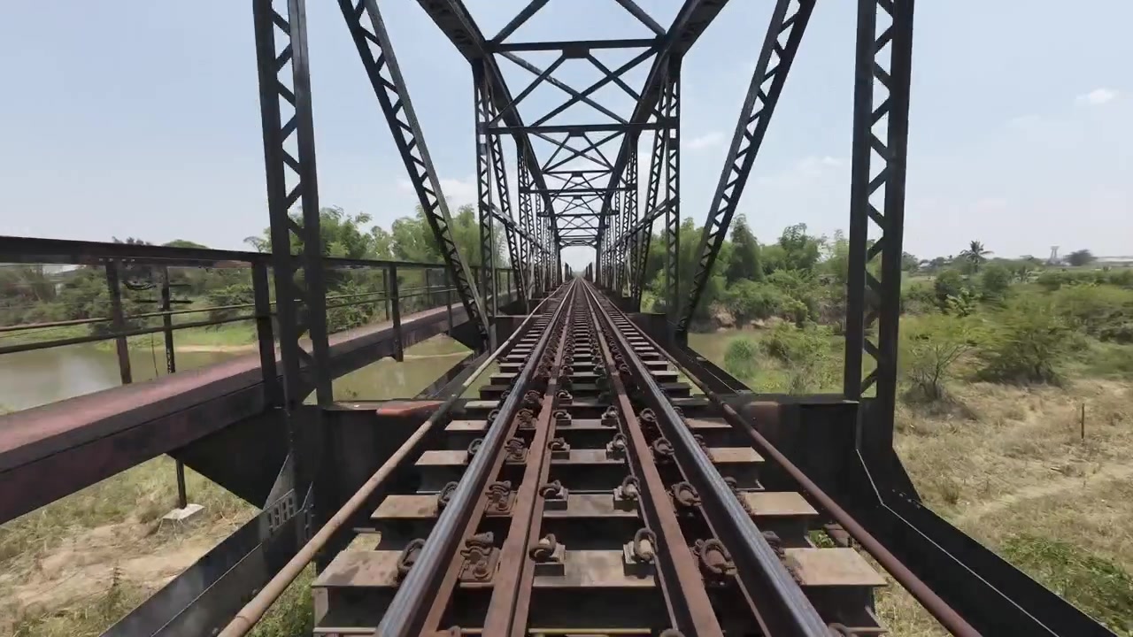 Vuelo FPV suave a lo largo de las vías del tren y el histórico puente de acero Tha Chang Black Bridge cruzando un río tropical en Nakhon Ratchasima, Tailandia