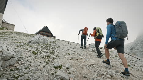 hikers with big backpacks going toward the mountain cottage climbing up on rocks and using hiking poles