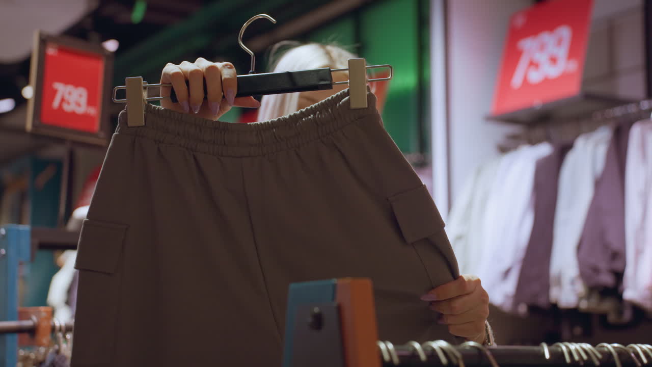 Stylish woman takes out baggy brown pant with side pocket feels it with her fingertips exploring texture and design under warm store lighting among organized racks of casual apparel