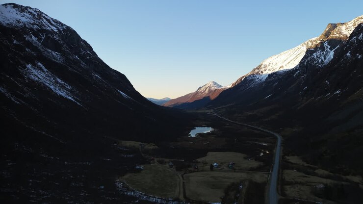Una strada si snoda attraverso una valle norvegese mentre le cime innevate catturano la luce dell'ora d'oro. Ideale per documentari di viaggio o sfondi panoramici