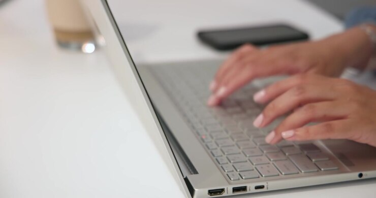 Donna africana che posiziona le mani e digita su un laptop a una scrivania bianca mentre finisce un compito, con telefono, tazza e smartwatch.
