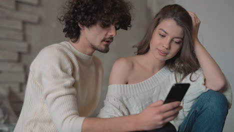 young cute couple sitting in bed looking something funny on the phone and wearing winter clothes