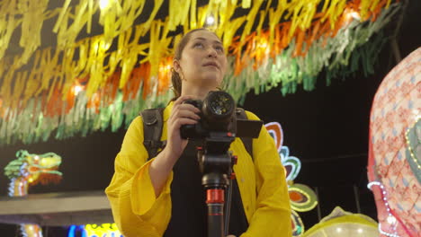 female photographer amazingly looking around at festive surroundings at night while holding on camera mounted on a tripod