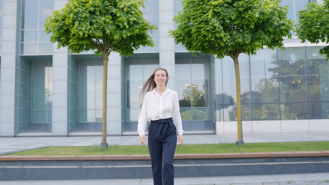 mujer de clase moderna caminando hacia la cámara cerca del edificio de oficinas, sonriendo