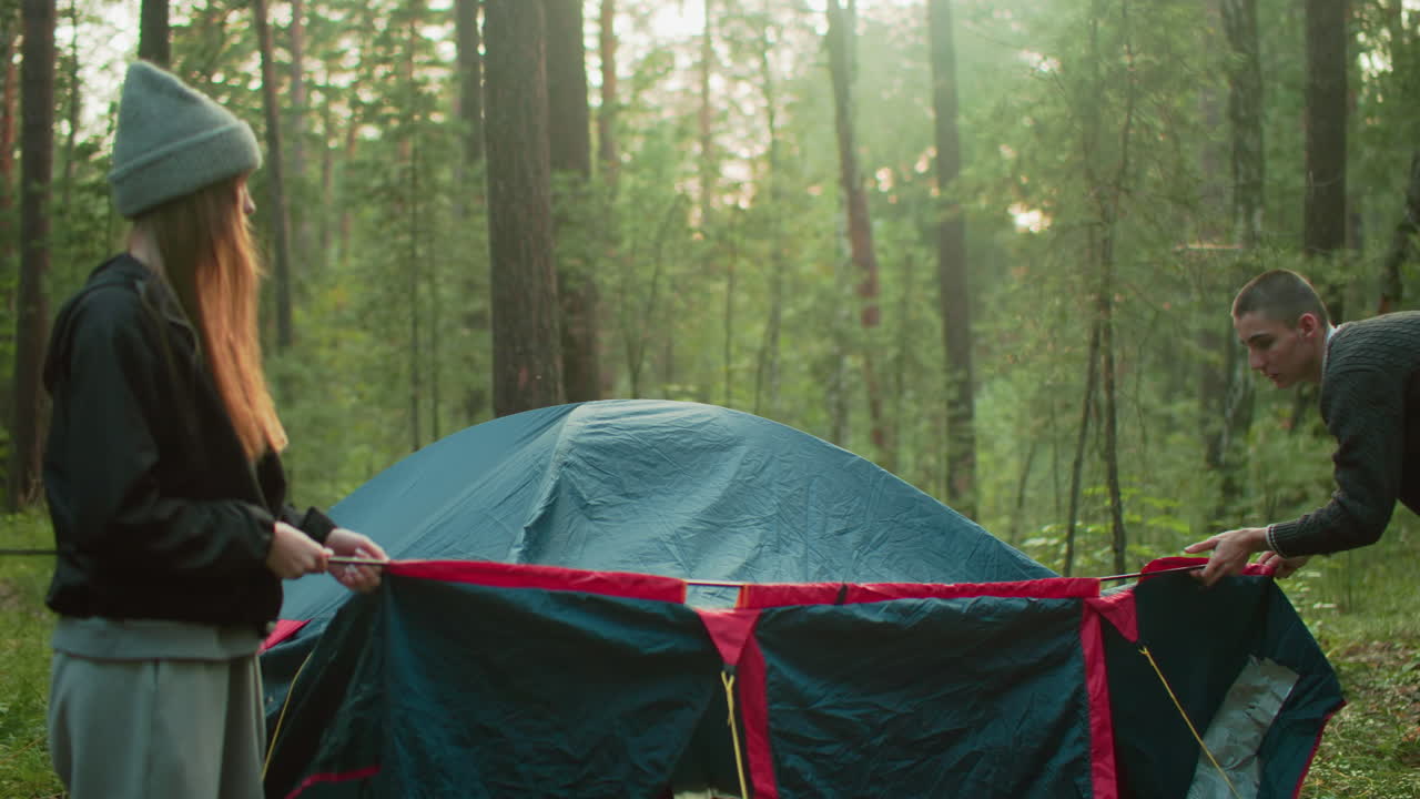 Lady holds iron pole steady as man carefully passes it through tent sleeve in forest, both engaged in collaborative camping setup surrounded by tall trees and soft daylight