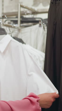hermosa dama de vestido rosa recupera la camisa blanca del estante de ropa, observándola de cerca, otras ropas están ordenadamente dispuestas en estantes y estantes en una tienda bien organizada, con un diseño elegante