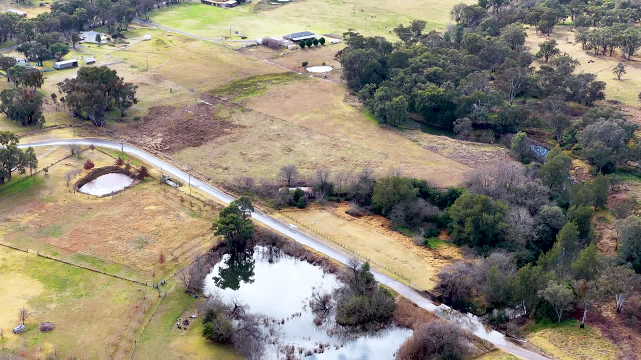 Drone footage glides above a winding rural road, ponds, and open farmland bordered by trees under soft daylight, revealing a peaceful countryside landscape