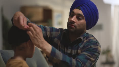 padre usando aguja salai para arreglar el cabello al atar el turbante a la cabeza del joven hijo sikh sentado en el sofá filmado en tiempo real