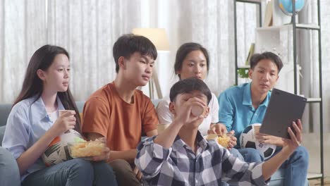 asian teenagers cheering, eating and drink while watching football game on tablet at home