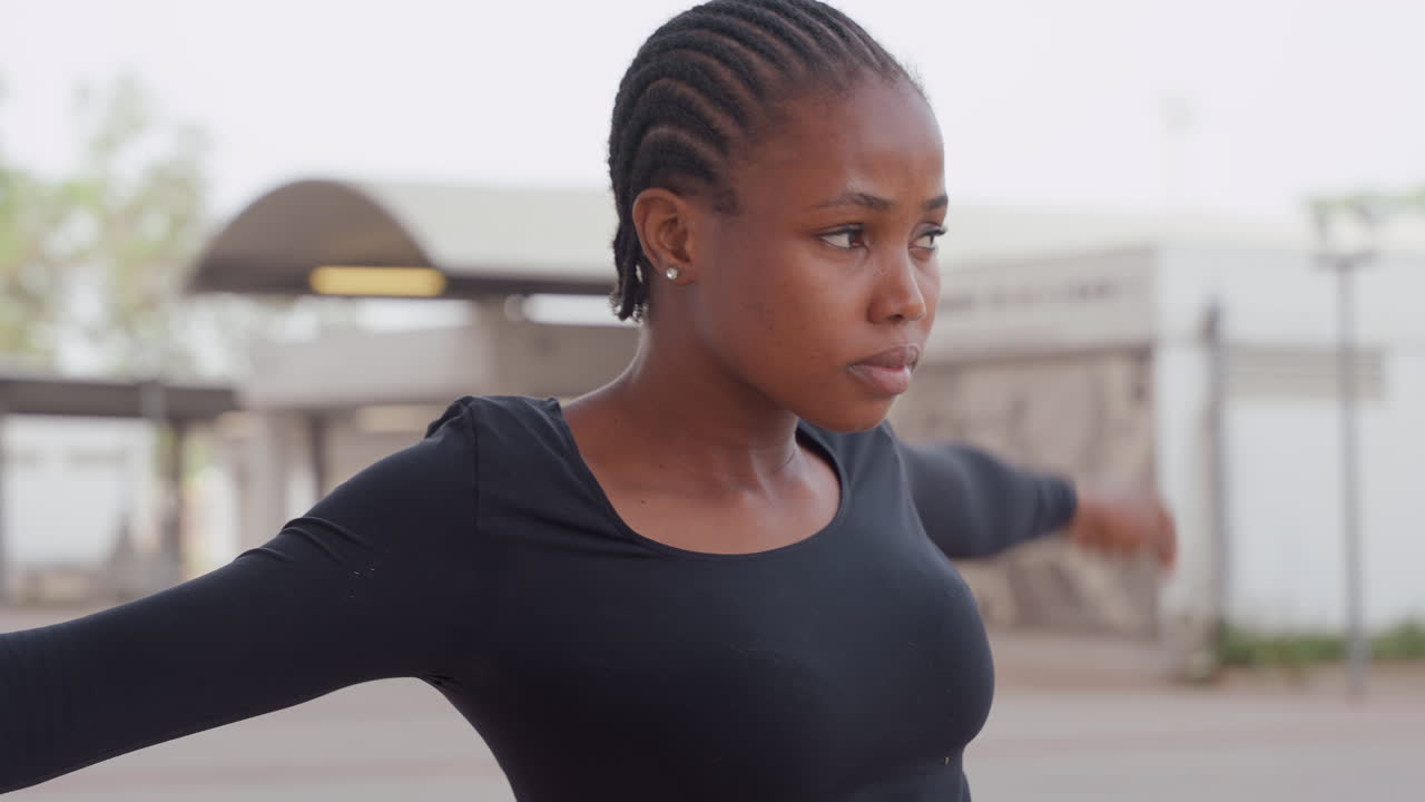 woman preparing for outdoor training, urban garage mural backdrop, arms extended for balance, determined gaze, pavement and canopy visible, candid fitness portrait with steady breathing