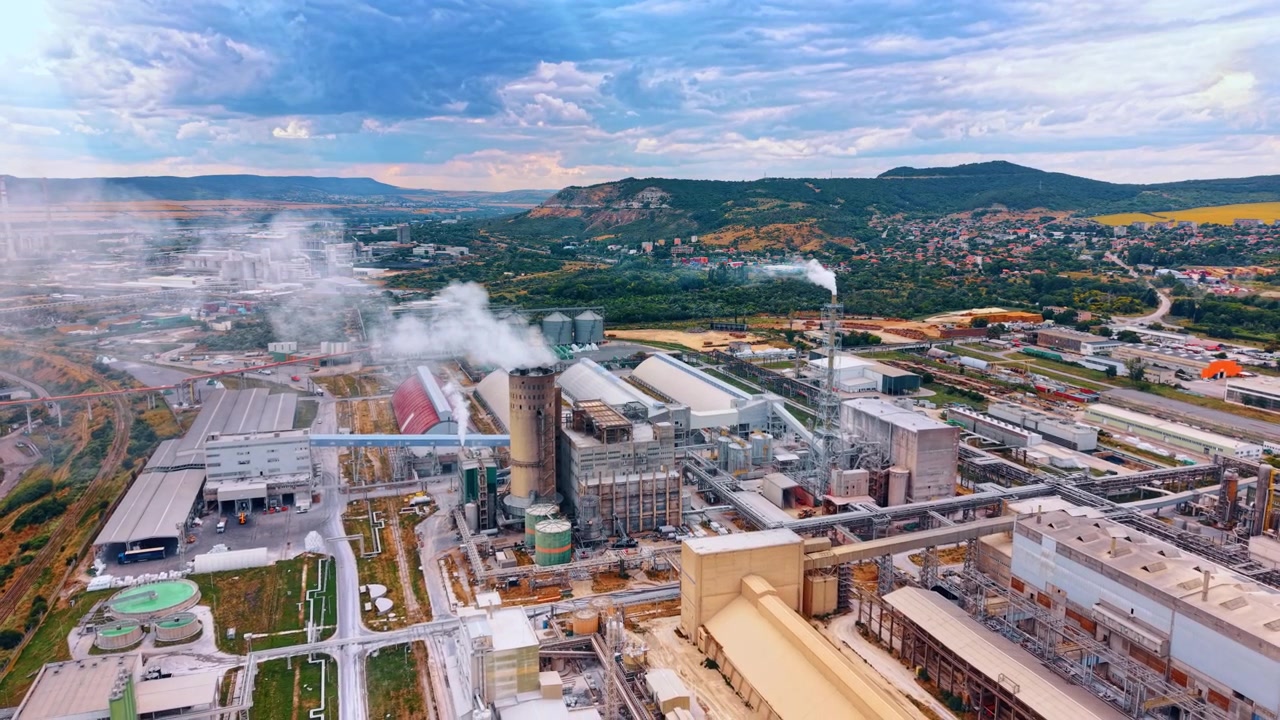 Steam rising from processing plant below hillside settlement. Factories, pipelines, chimneys and storage silos fill a valley beneath dense cloud cover.