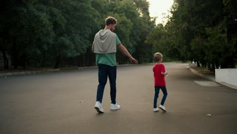 Rear-view-of-a-dad-with-curly-hair-in-a-green-T-shirt-and-blue-jeans-and-his-little-blond-son-in-a-red-T-shirt-and-blue-jeans-walking-along-a-spacious-road-in-a-green-park