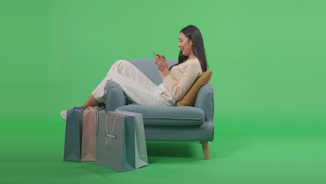 an asian shopping woman with shopping bags enjoy using mobile phone shopping online while sitting on sofa in front of green screen