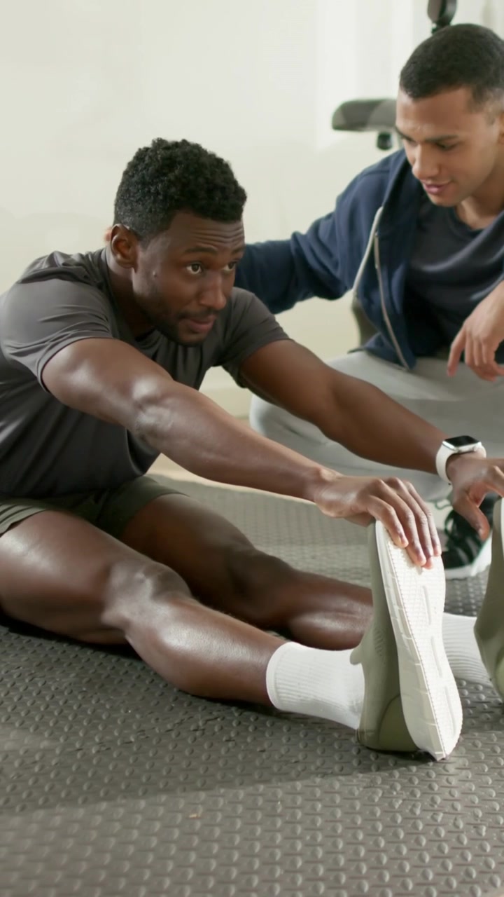 Vertical video: After cue man gray tee reaching for shoe on gym mat for mobility coach with watch
