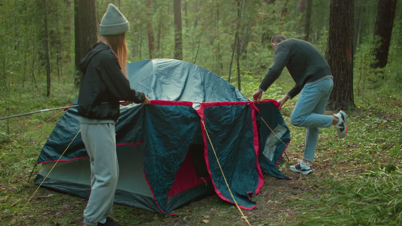 Couples working together to prepare tent in forest, holding flexible poles and adjusting tent cover, surrounded by greenery and trees, dressed in casual clothing