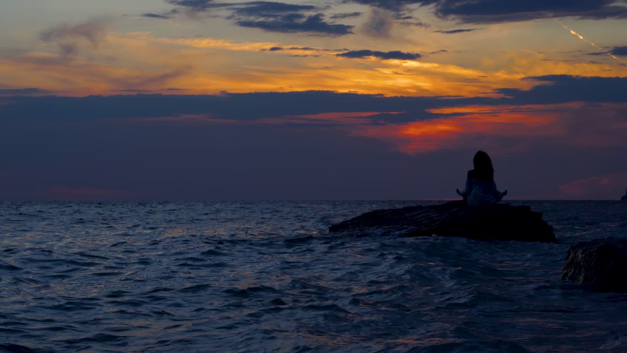 mujer sentada en una roca rodeada de agua de mar oscura al hermoso atardecer, haciendo ejercicios de yoga