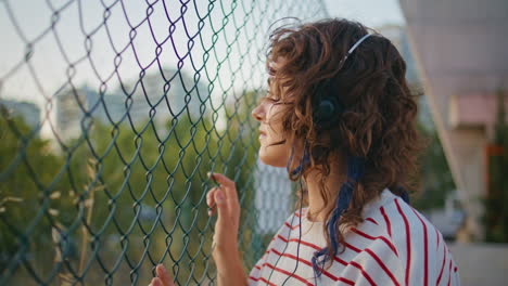 adolescente rizado escuchando auriculares de música mirando la valla de malla al aire libre de cerca.