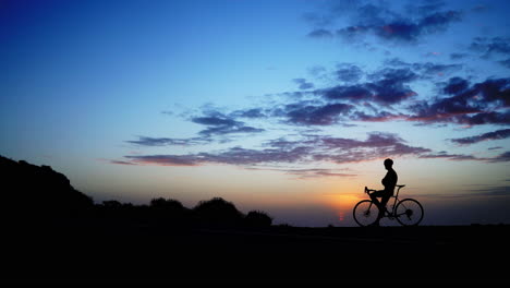 the man is standing with a bicycle set against the backdrop of a sunset, captured in a time-lapse sequence. the scene is captured from a wide-angle perspective