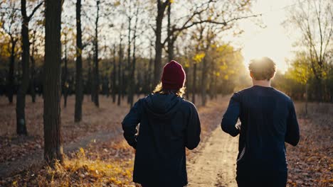 Rear-view-of-two-male-athletes-in-a-black-brunette-sports-uniform-running-along-an-earthen-path-in-a-sunny-autumn-forest-during-their-jog-in-the-morning