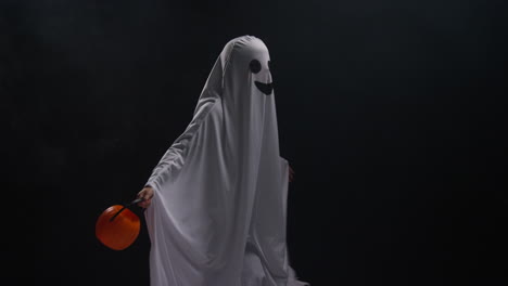 Child-Dressed-Up-In-Ghost-Costume-Trick-Or-Treating-At-Halloween-Holding-Pumpkin-Shaped-Jack-O'Lantern-Bucket-Against-Black-Background-1