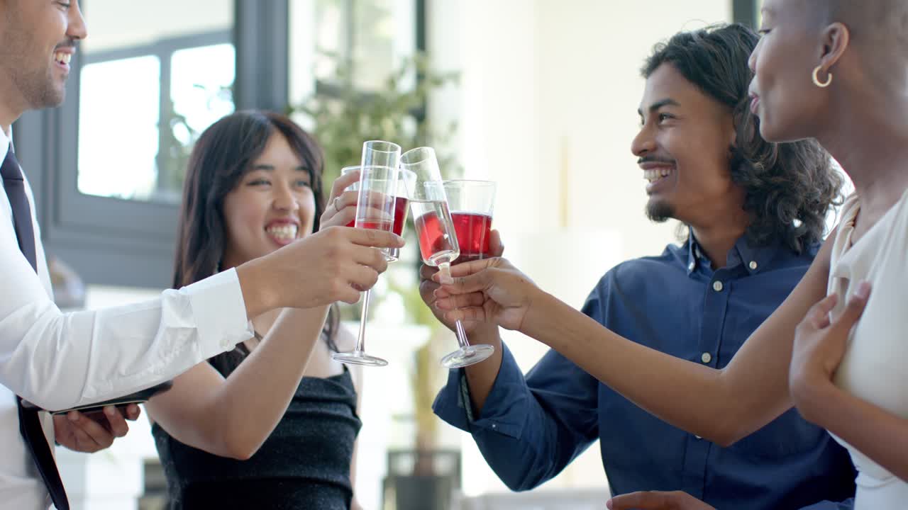Diverse friends in formal attire smiling in bright lounge, raising flutes and tumblers for toast