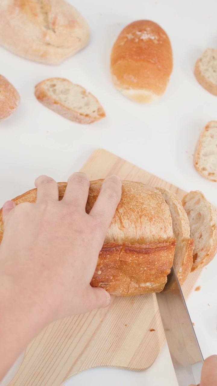 Hands slice a crusty sourdough loaf on a wooden board against a white background.
