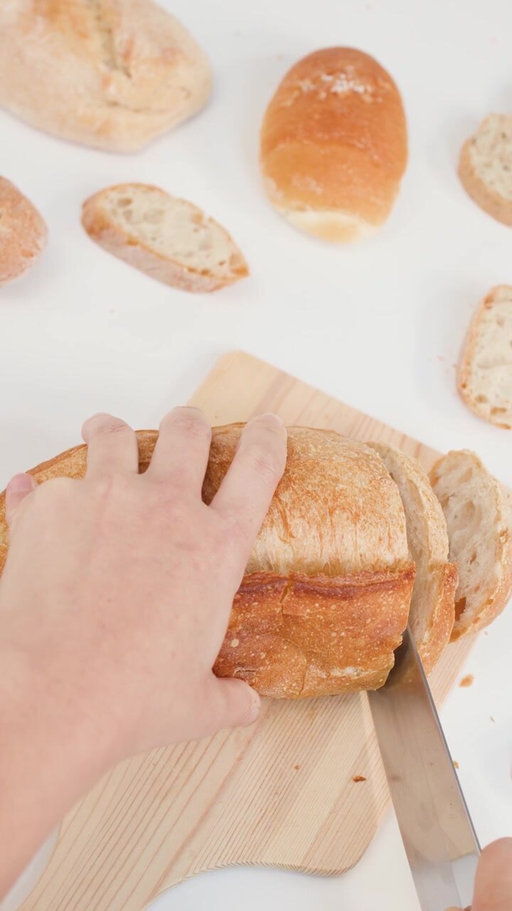 Hands slice a crusty sourdough loaf on a wooden board against a white background.