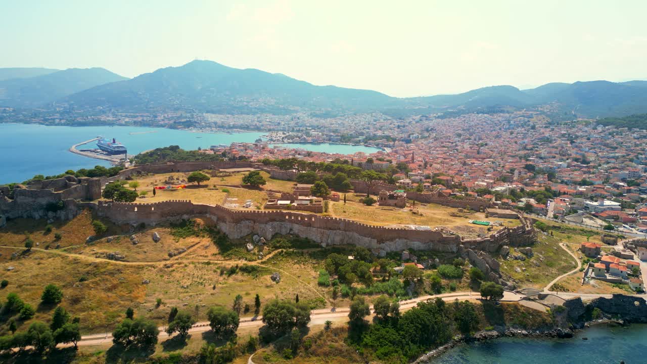 Wide aerial shot orbiting the Castle of Mytilene, Greece, with a backdrop view of the port, terracotta roofed houses, and mountain hills.