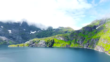 aerial drone footage flying over a large blue lake that has steep large mountains with cliffs with green moss on them during a sunny day in lofoten norway. large white clouds and mountains visible.