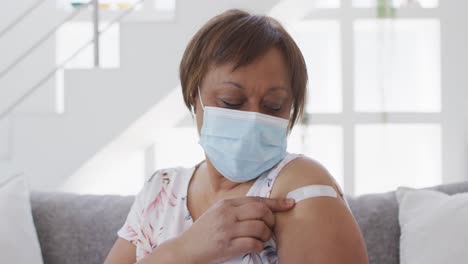 african american senior woman in face mask showing bandage on arm after covid vaccination