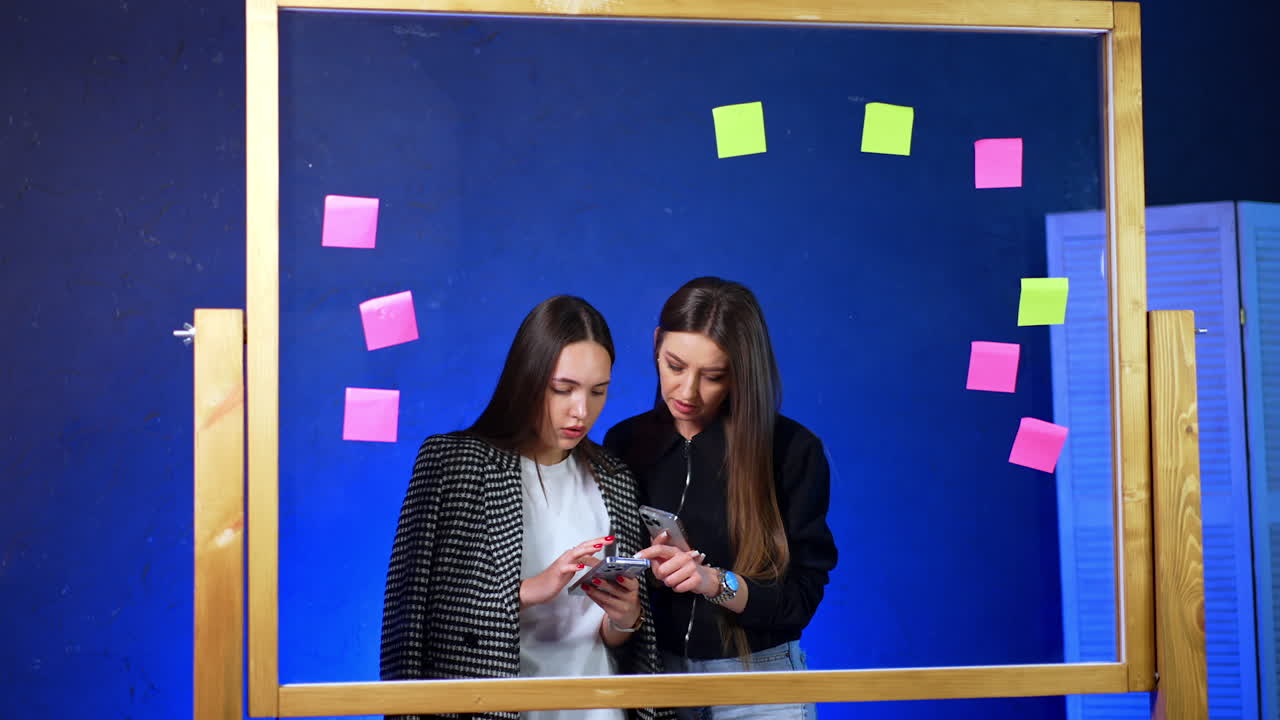 Friends using phones in space. Two young women engage with their smartphones, surrounded by colorful sticky notes in a modern workspace