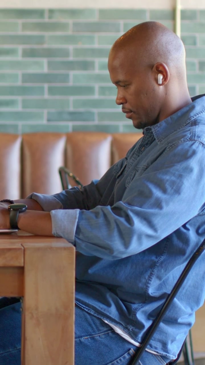 Vertical video: Settling into chair man leaning forward and working at table at cafe, with earbuds