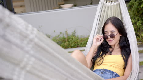 a young biracial woman lounges in hammock with sunglasses on, copy space