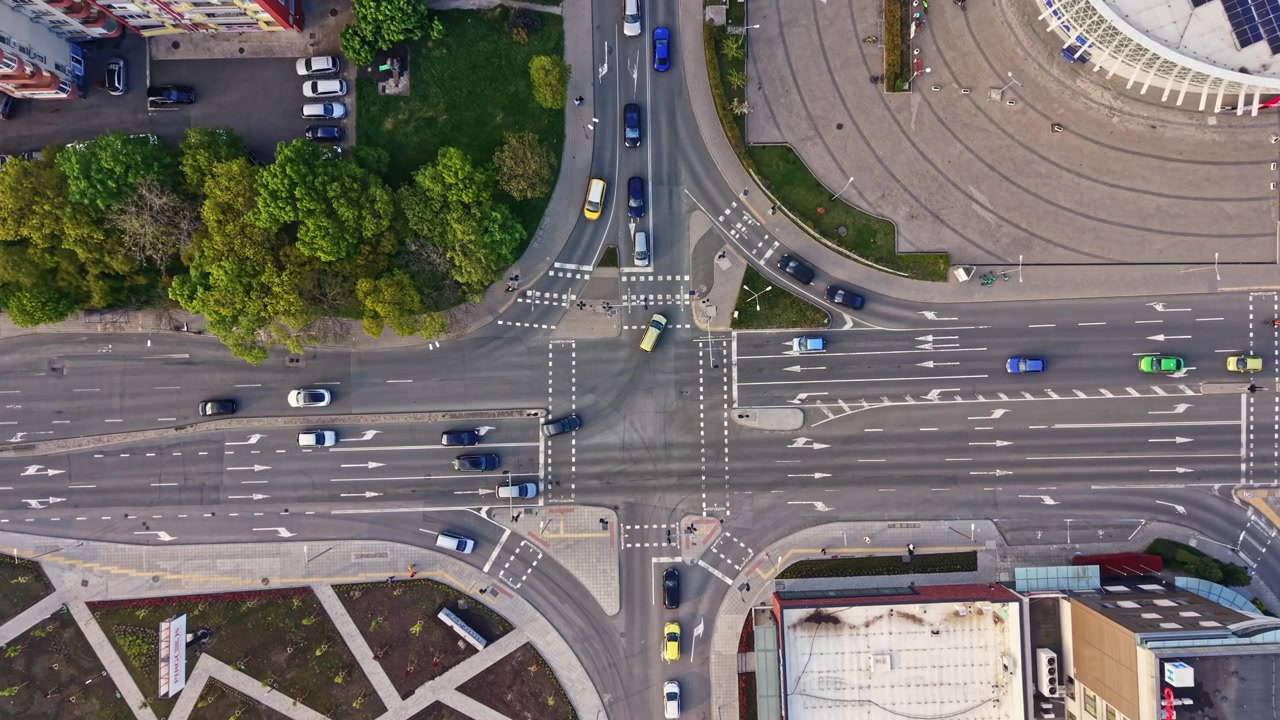 Traffic flows through an intersection in an urban area during the day with multiple vehicles and trees lining the roads