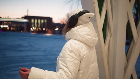 Back view of creative wearing white hoodie coat and headphones, rhythmically moving her body inside lighted pavilion in snow covered park during sunset glow, with city lights and bare trees
