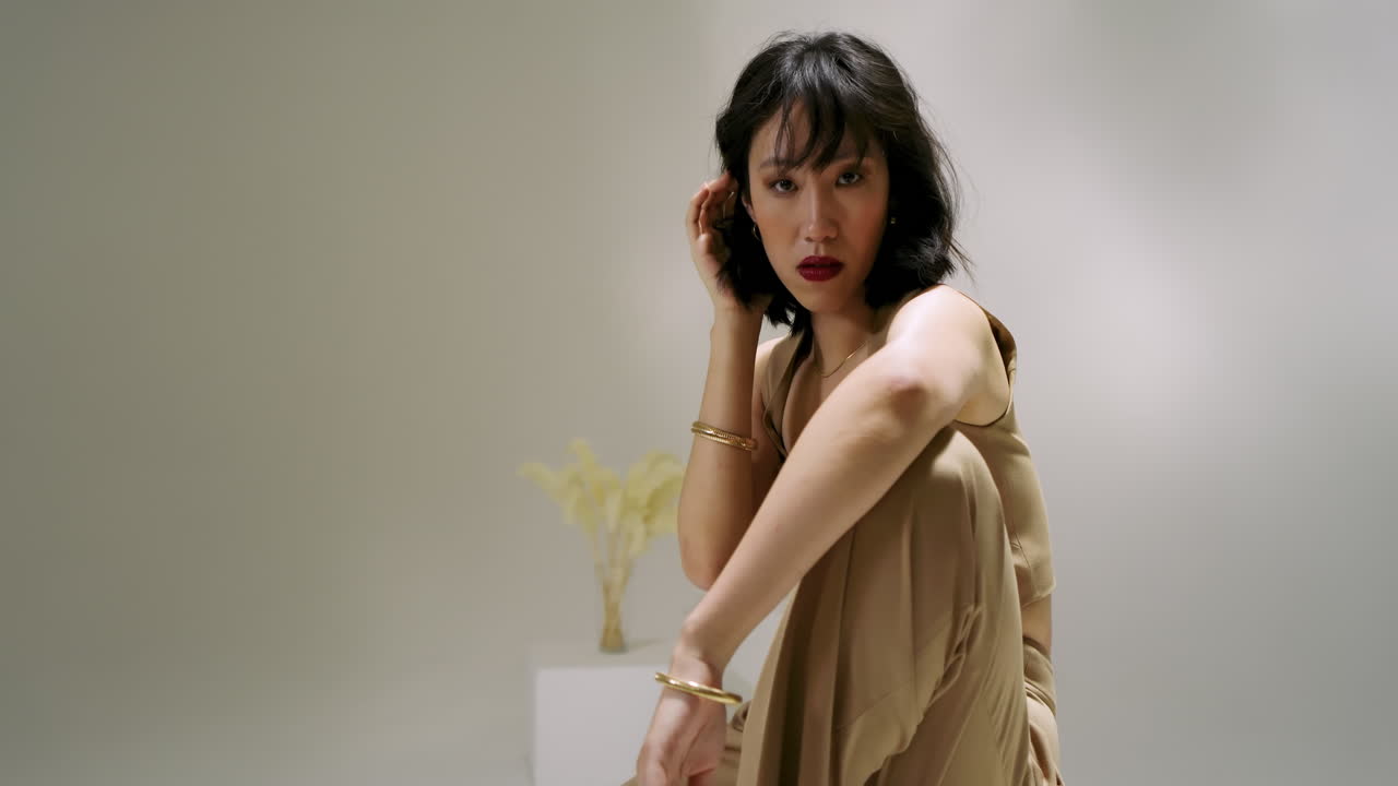 Upon start Chinese woman lowering onto white cube in studio, adjusting gold bangle for photoshoot