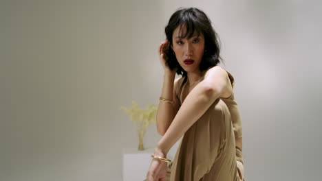 upon start chinese woman lowering onto white cube in studio, adjusting gold bangle for photoshoot