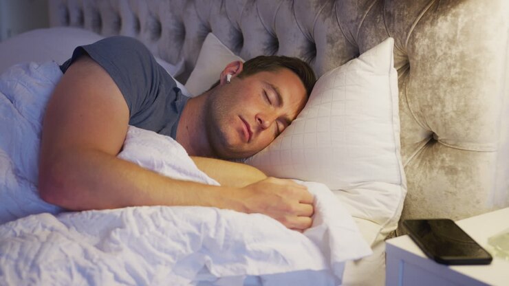 Man Sleeping In Bed Wearing Wireless Earphones Connected To Mobile Phone On Bedside Table