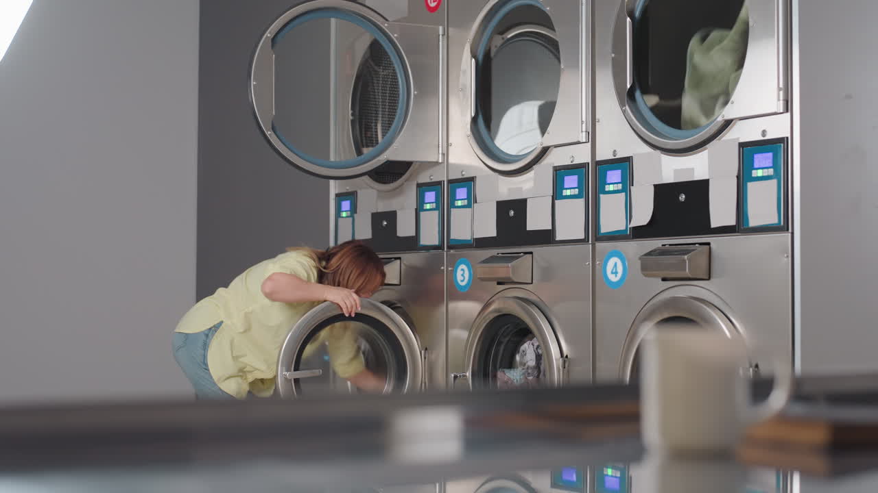 Female manager transferring clothes from washer into dryer inside laundromat, leaning forward with focus while handling laundry tasks, stainless steel machines with digital panels