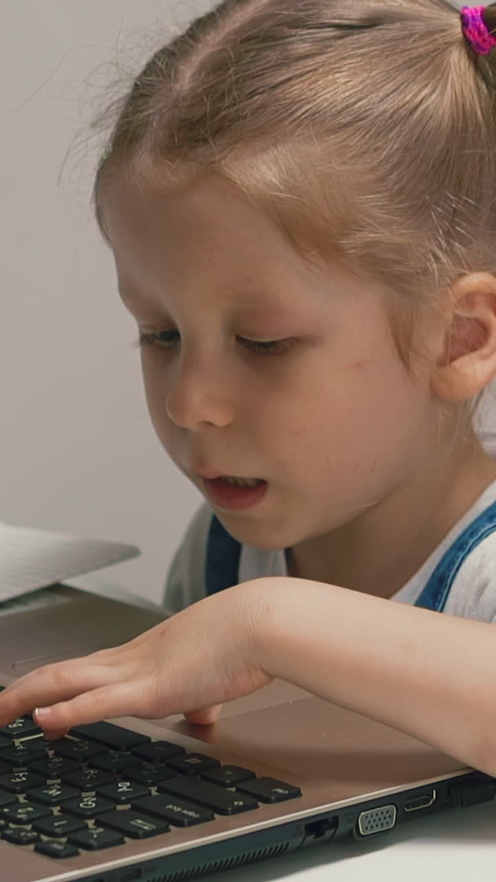 Slow motion: adorable little girl in white t-shirt with blonde plaits plays on modern laptop sitting at table in brightly lit room at quarantine time